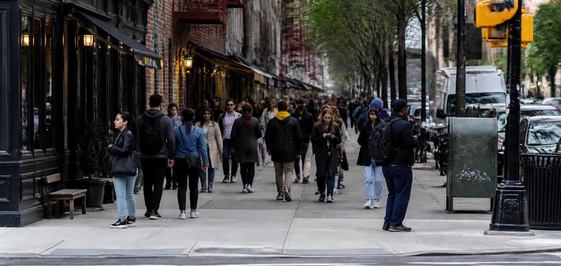 Wide view of a dense New York City sidewalk with pedestrians walking past storefronts