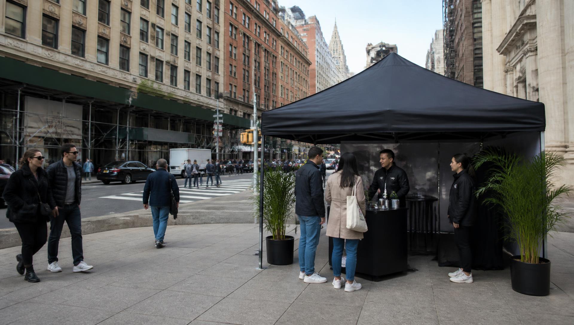 A street-level brand activation tent on a wide New York City plaza with pedestrians passing and a team talking with people